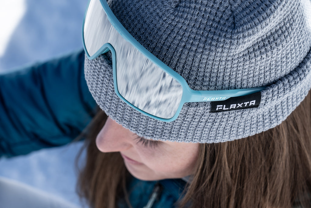 Person wearing a gray knit beanie with blue rimmed goggles, featuring the Flaxta brand logo.