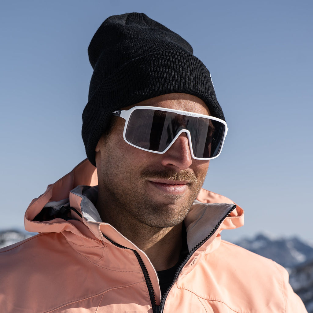 Person wearing a black beanie and peach jacket with the Above sunglasses in black and white against a mountainous background